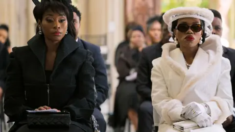 Two women seated next to each other at a funeral. The one of the left in a smart black outfit and hat, the other on the right in a fashionable white outfit with fur trim and white gloves.