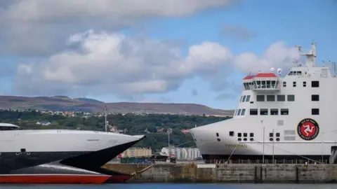 The bows of the Manannan and the Manxman, the Manannan, a sleek catemaran with black, white and red, faces the Manxman a tall boxy ferry, they are both at Douglas harbour.