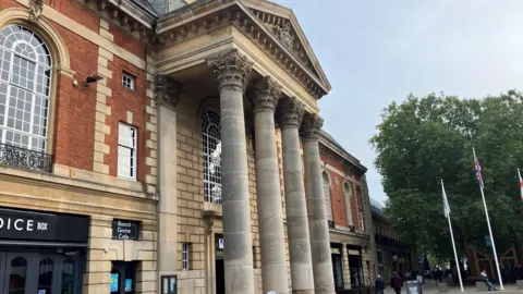 Outside of Peterborough town hall, where there are four large columns in front of the brick building in the city centre.