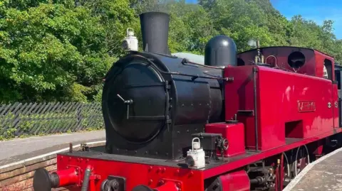 Peak Rail A red steam engine with a black front and chimney. It is parked up on a station platform surrounded by woodland.