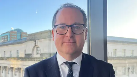 Claudia Savage/PA Wire Donal Lunny, chair of the Bar Council of Northern Ireland, posing for a photo in front of a large window overlooking Belfast City Hall. He is a man with short, dark hair and dark-rimmed glasses. He is wearing a navy suit and tie with a white shirt. 
