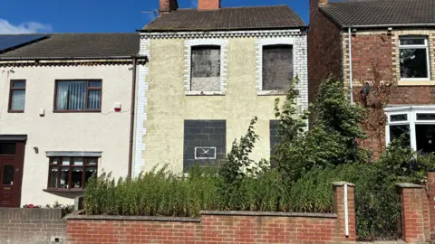 A boarded up semi-detached house with an overgrown front yard. The weeds have grown above the wall around the perimeter of the property and large trees are bending in the wind.