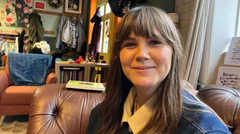 Vikki Irwin/BBC This shows a cozy indoor setting in cafe with leather armchairs with a woman sitting on one. She has long dark hair and is wearing a white collared shirt. In the background there is patterned rug, and a wall decorated with coats, framed pictures, and shelves.
