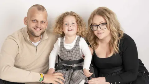 Tony and Carrie Pearson sitting outside with their daughter Grayce. Grayce is smiling widely at the camera and has shoulder-length, curly, blonde hair. She is wearing a white, long-sleeved top and a brown checked pinafore dress. Carrie is wearing a black top and skirt and has long, blonde hair and black-rimmed glasses. Tony has a shaved/bald head and a beard and is wearing a beige, long-sleeved top.