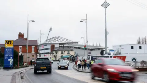 PA Media The Stok Cae Ras as seen from Mold Road in Wrexham, with the Turf pub also seen to the side of the stadium, with a teal-coloured gazebo seen next to it. Some people are walking on the pavements to either sides of the road and several cars can also be seen on the road.