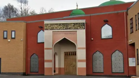 Google An exterior shot of Oldham Central Mosque- the building - part red brick and part beige brick- has mosaic windows and green domes. Oldhanm Central Majhid is written on the door