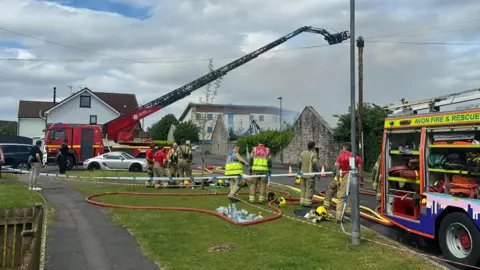 Several fire engines, including one with a turntable ladder, parked in a residential area. Several crew members can be seen standing on a grass verge beside the road and there is smoke in the air, coming out of a derelict building.