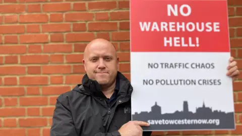 Laura Coffey/BBC A man wearing a black jacket and holding a red and white sign saying "No Warehouse Hell - no traffic chaos, no pollution crises, save Towcester now". He is standing in front of a brick wall.