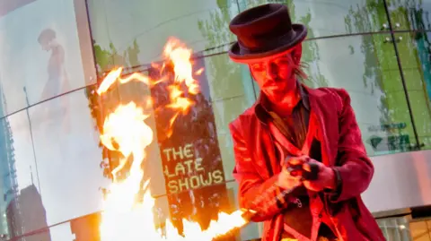 The Late Shows A man in a black top hat and red coat and waistcoat holding a silver chain which is on fire. He is looking down at it. Behind him there is a glass wall with a black sign which reads 'The Late Shows' in yellow writing