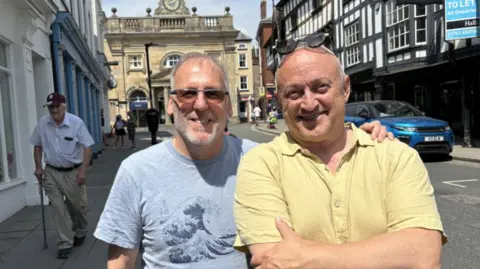 Two men with grey hair and sunglasses, one withe a pale blue top and the other with a pale yellow top, standing in a busy street with timber-framed buildings and an ornate stone building in the distance
