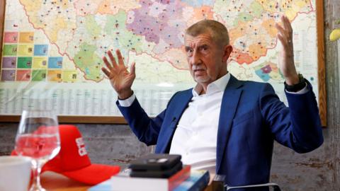 Andrej Babis gestures with both arms lifted, sitting in an office with a map of the Czech Republic behind him. There is a metallic-looking cylindrical vase of white flowers to the right of the picture. On the table in front of him are a red hat, a wine glass half full with clear liquid, and a white teacup. Babis is wearing a white shirt and navy blue jacket. He has grey stubble and short, cropped greying hair.