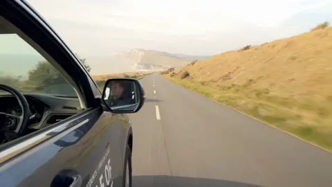 A man is seen in the side mirror of a car driving down a road with a grassy bank on the side. There are cliffs in the distance. The car is blue and the window is down.