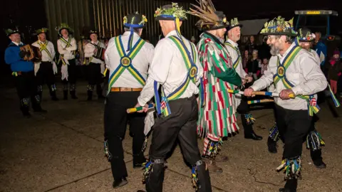 Tu Plus Tu Photography Morris dancers circle around Simon in the farmyard. They wear white shirts, black trousers and bells around their knees, and hold coloured wooden sticks. To the side is a man with an accordian.