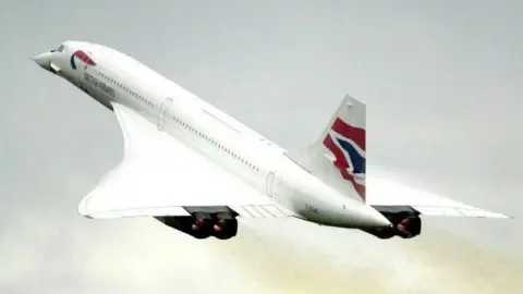 PA Media A Concorde plane flying in the sky. It is a white plane with wide wings and a pointed droop nose at the front, under the cockpit windows. It has red and blue British Airways branding on it. 