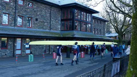 Ian Gordon People walking while carrying a long yellow rowing boat on their shoulders. They are in shorts. In the background is a large brick building with windows. Onlookers walk past. 