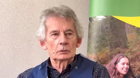 Martin Heath/BBC John English with medium-length white hair wearing a blue shirt with white spots and a blue waistcoat. He is sitting in front of a poster which has picture of a young woman with long brown hair standing in front of a tree.
