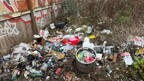 LDRS Lots of plastic bottles, a barrel and food packets strewn about next to a heavily graffitied fence. 