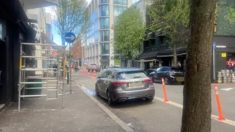 A grey Mercedes parked inside a mandatory cycle lane in Belfast. Orange bollards are on the right of it. Buildings are all around.