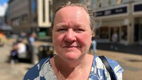 Debbie O'Loughlin is white and has a blue and white dress, a gold necklace and tied back hair