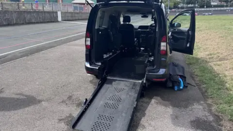 A black long car, specially made for disabled users, with a black metallic ramp coming out at the rear, parked on the side of the road.