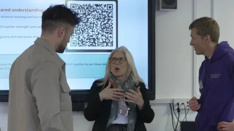 Good Future Foundation Stoke-on-Trent South MP Allison Gardner, wearing glasses and a black suit jacket, is stood talking to two men, one is wearing a light-coloured jacket and the other is wearing a purple hoodie.