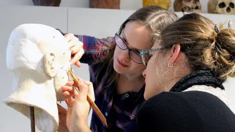 Two people with long hair and glasses are both working on the sculpture of a face with various tools. 