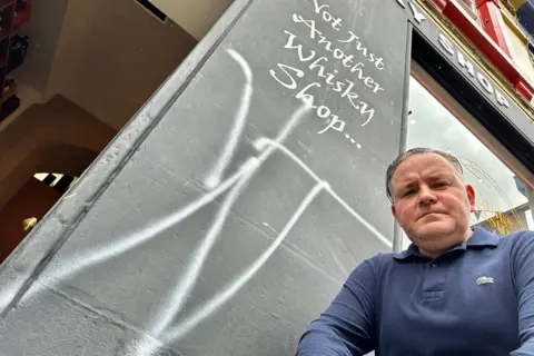Andrew is looking down at the camera. He is wearing a blue top. There is white graffiti beside him on a shop.