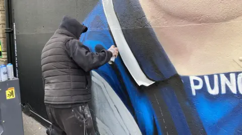 Steve Martin/BBC A gentleman wearing a black hoodie, black cap and black puffer jacket is pictured spray-painting the mural. He is painting the collar of Sheeran's football top on the mural.