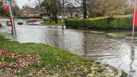 A residential street that has flood water completely covering it. There is a bus stop sign on the left, and grass next to it. There is a black car driving towards the water in the distance