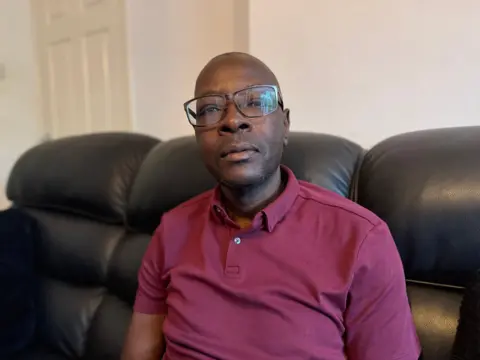 Maxwell is sat on a leather sofa and is wearing glasses and a burgundy t-shirt. 