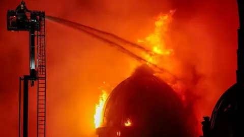 Getty Images A huge fire engulfs the dome of the building. Firefighters are dousing it in water from hoses.