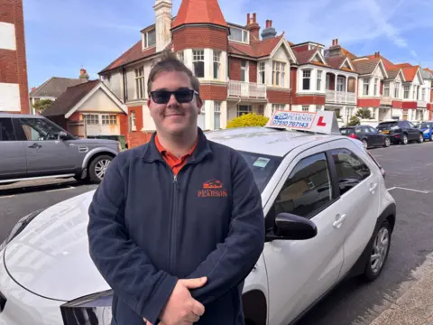 Driving instructor Joe Pearson, wearing sunglasses, smiles as he stands in front of a car.