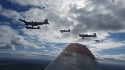 Tony Blake  A view from the plane, and the planes fly in formation over Gloucestershire. The wing of the plane the photographer is in can be seen, and five light aircraft are in the sky. The sky is blue with lots of white clouds.