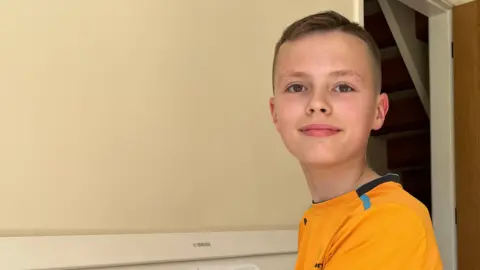 Harvey, 11, sits at a white piano with his hands on the keys and looks at the camera. He is wearing a gold-coloured replica F1 shirt. 
