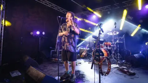 Dom Mosley  A man in a shirt standing on stage in front of a microphone and drum kit