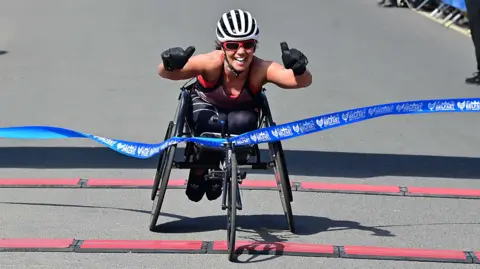 A lady in a wheelchair with a white helmet puts her thumbs up as she crosses through the blue ribbon. She is wearing red glasses and is wearing black gloves. She is wearing a vest with leggings.