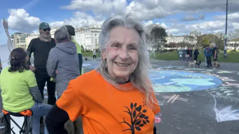 Ann Read smiling at the camera in front of the Earth artwork. She is wearing an orange top. 