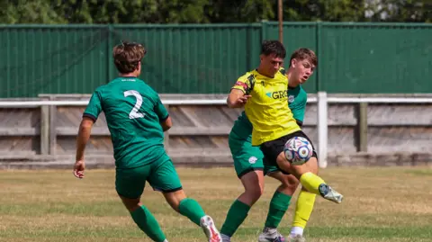 Button Oak Photography A football action photo, featuring a pitch showing little green grass from this angle. Two players, one in a green shirt and another in a yellow shirt, are challenging for the ball. A green fence is in the background.