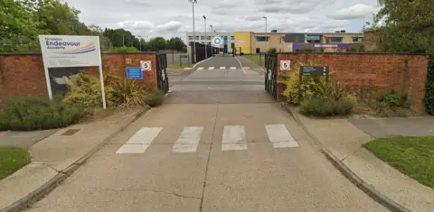Google Front of Ormiston Endeavour Academy with a sign on left side of a wall with a road going through a gate. 