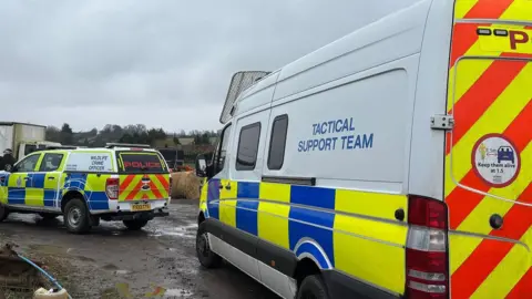 Cambridgeshire Police Two police vehicles parked. One police vehicle says POLICE in red writing on the back. The other is a larger van which reads TACTICAL SUPPORT TEAM. 