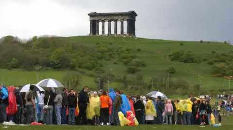 A crowd of people wearing waterproof ponchos and holding umbrellas stand in a queue in a field. On a hilltop beyond them is the Penshaw Monument, a memorial made of large stone columns.