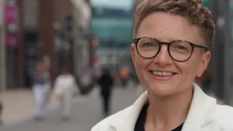 Labour Party Bev Craig - a woman with short red hair, wearing glasses and a white jacket over a black blouse. She wears a Manchester Bee badge on her jacket lapel.
