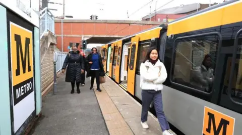 Nexus Passengers walk along a Metro platform at Millfield station next to the new train. It has a yellow-and-grey livery with the Metro logo on the side of one of the carriages.