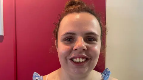 A picture of Julie Griffin taken against a pink backdrop, she's wearing her brown hair in a bun and is smiling.