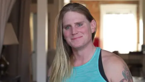 Kara's long straight blonde hair falls over her right shoulder. She is wearing a light blue vest top and has a tattoo on her left arm.