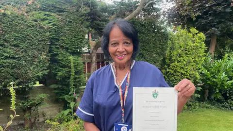 Sofía Luis-Hobbs/BBC A woman in purple scrubs smiles at the camera as she holds up a certificate in front of her. She wears her black hair up and has a multi-coloured striped NHS lanyard around her neck as she stands in a garden
