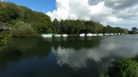 River bank with shrubs bridge over the water to left and boats along a far side bank lined with trees