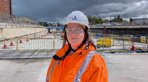 A woman wearing a bright orange hi-vis jacket and white safety helmet, She is standing in an area that is under the level of a main road with cones and barriers in the background.