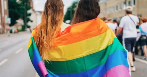 Getty Images Two people stand with their back to the camera, wrapped in a rainbow flag as a parade proceeds down a street