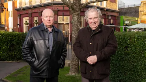 PA Media Paul Bradley as Nigel Bates (right) wearing a brown coat with this hands clasped in front of him, standing next to Steve McFadden as Phil Mitchell, who is wearing a black leather jacket with his hands in his pockets, in Eastenders.